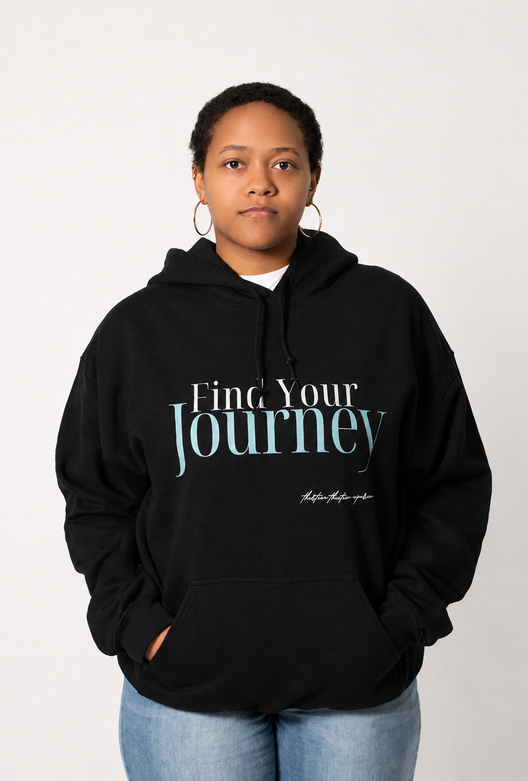 A person wearing a black pullover hoodie with the text 'Find Your Journey' printed on the chest, paired with light blue jeans, against a plain light gray background.