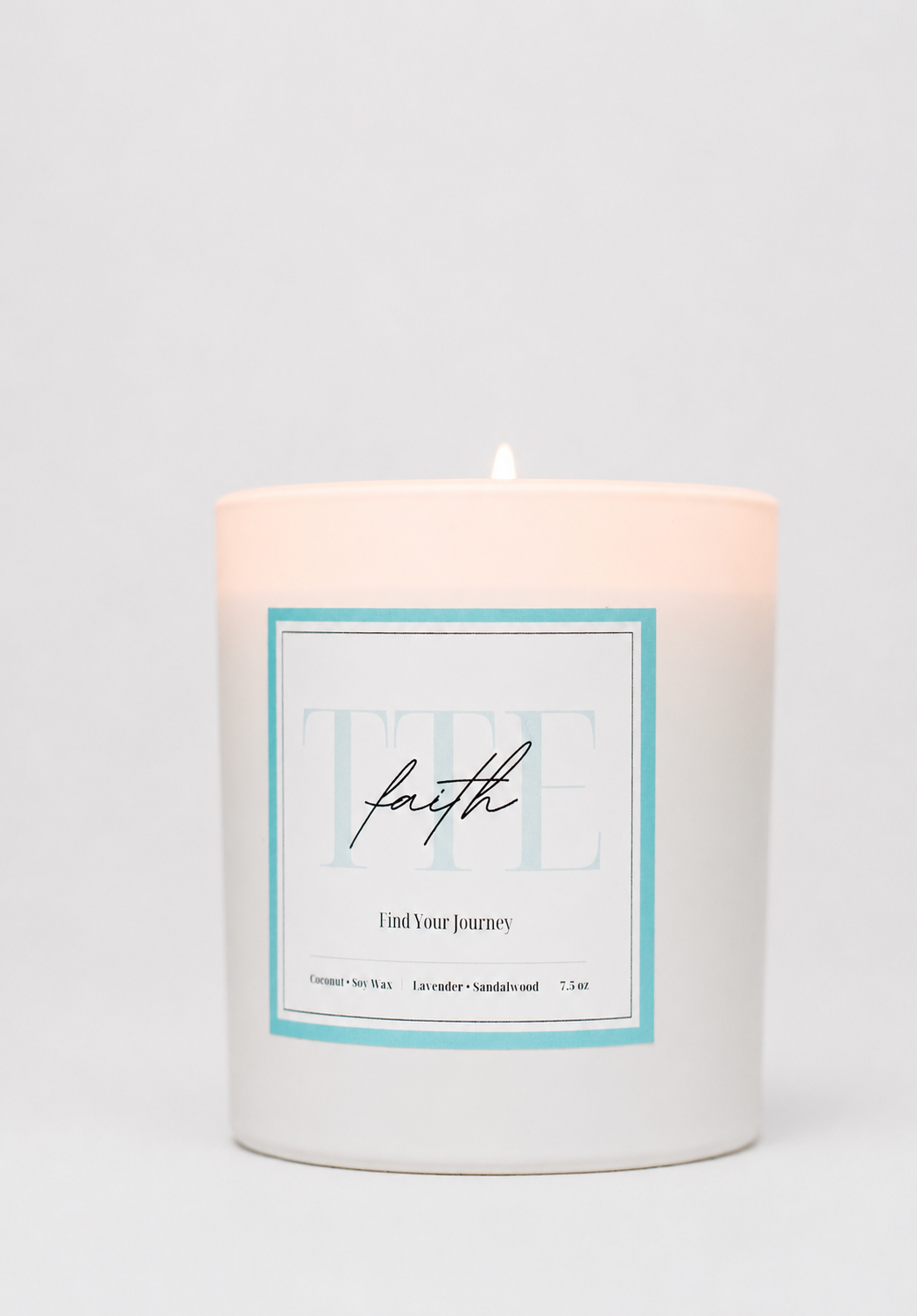 A white wax candle in a cylindrical glass jar with a lit wick, featuring a front label with a light blue border and the word 'faith' in script, set against a plain light gray background.