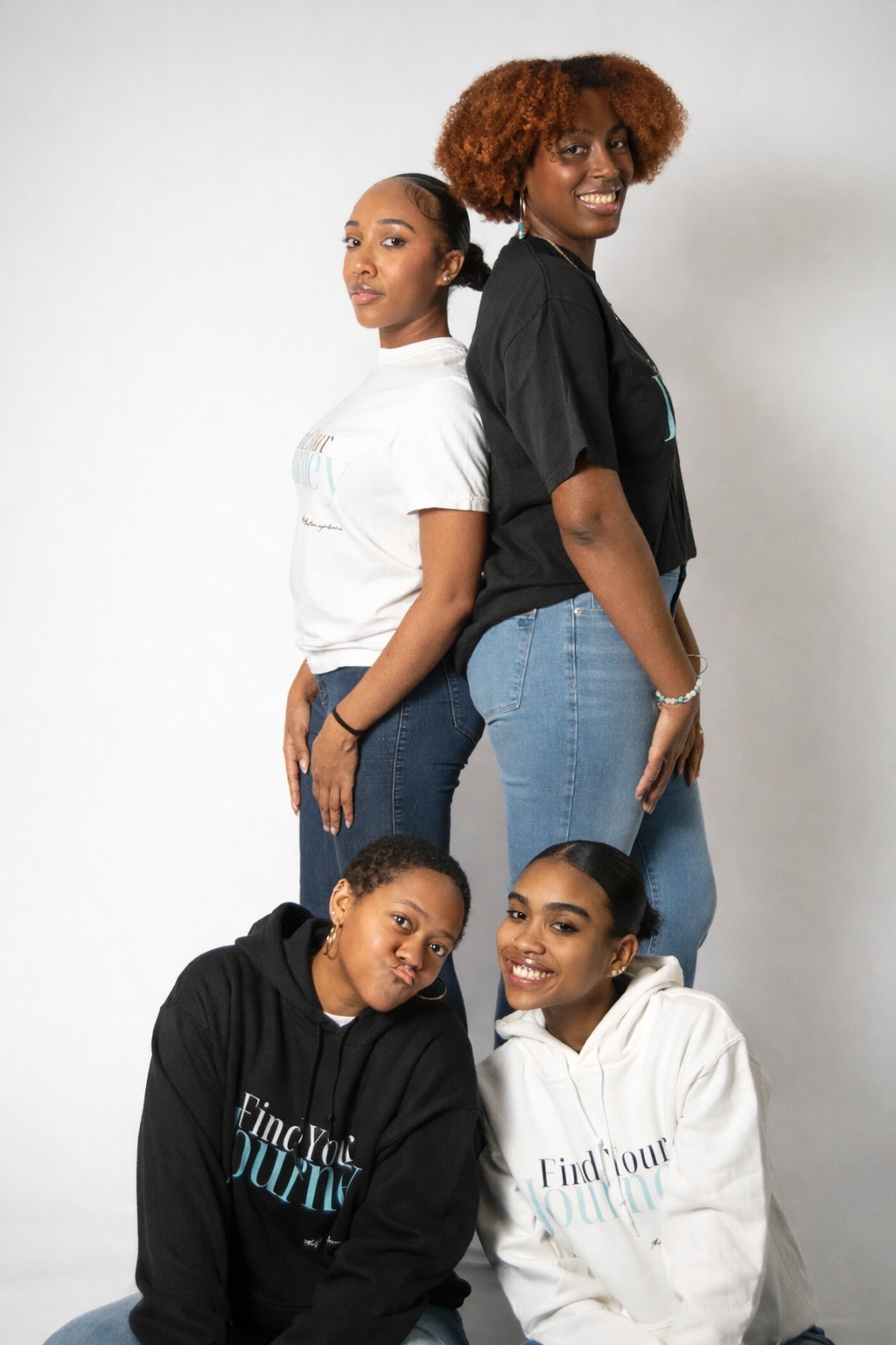 Four young women posing together against a plain light gray studio background. Two are standing and two are sitting, wearing casual hoodies and t-shirts in black and white with light blue text reading 'Find Your JIU JITSU'.