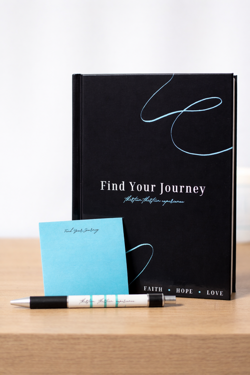 A black hardcover journal titled 'Find Your Journey' with white and light blue text and line art, displayed on a wooden surface next to a light blue sticky note and a black-and-white pen.