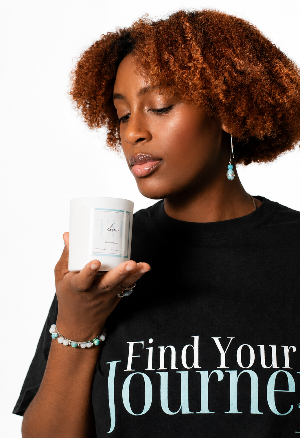 A woman with curly brown hair holds a white candle jar near her face. She is wearing a black graphic T-shirt that reads 'Find Your Journey' in white and light blue text, along with blue earrings and a beaded bracelet, against a plain white background.