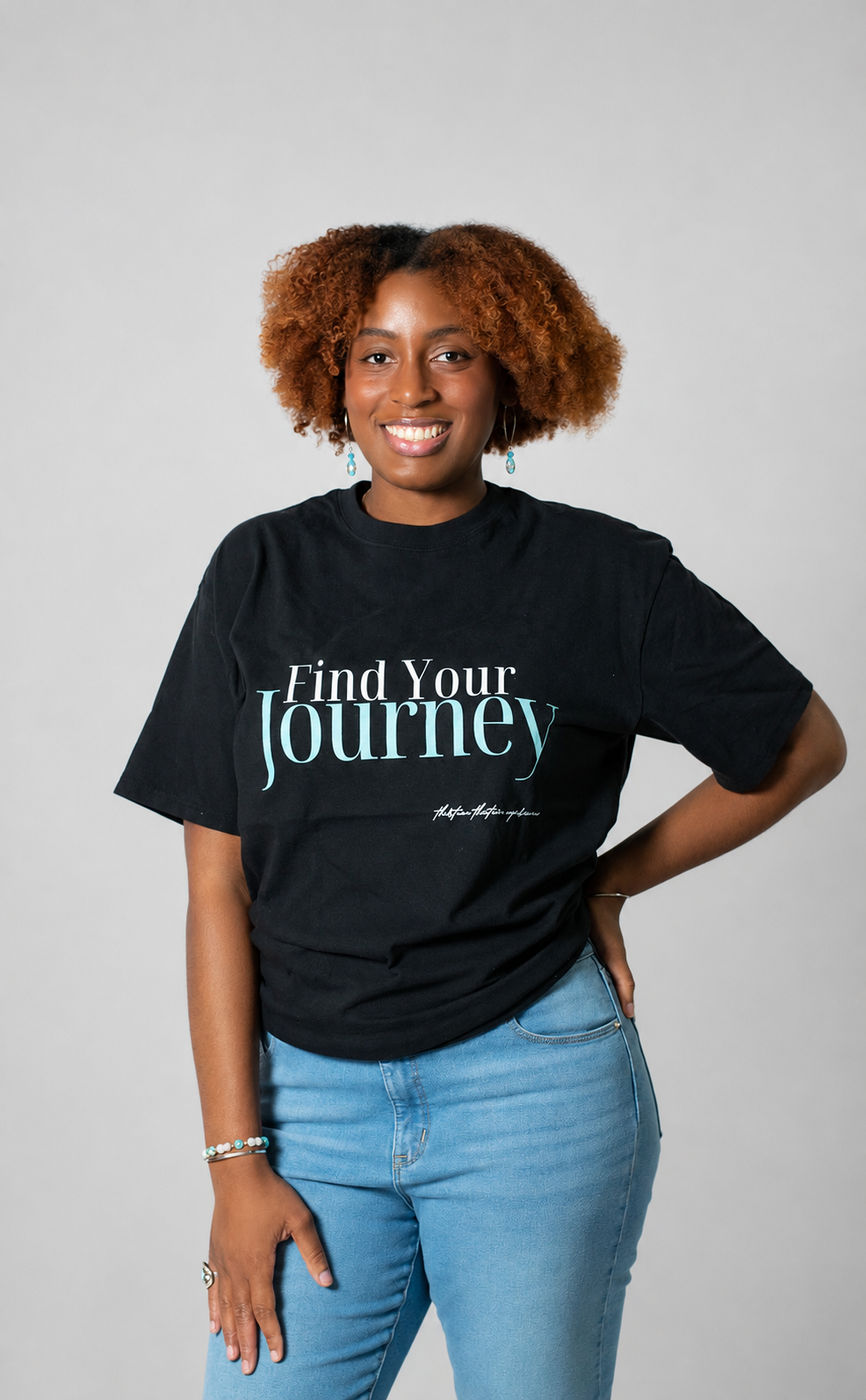 A woman with curly brown hair wearing a black graphic t-shirt that reads 'Find Your Journey' in white and teal text, paired with light blue jeans, standing against a solid gray studio background.