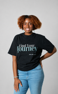 A woman with curly brown hair wearing a black graphic t-shirt that reads 'Find Your Journey' in white and teal text, paired with light blue jeans, standing against a solid gray studio background.