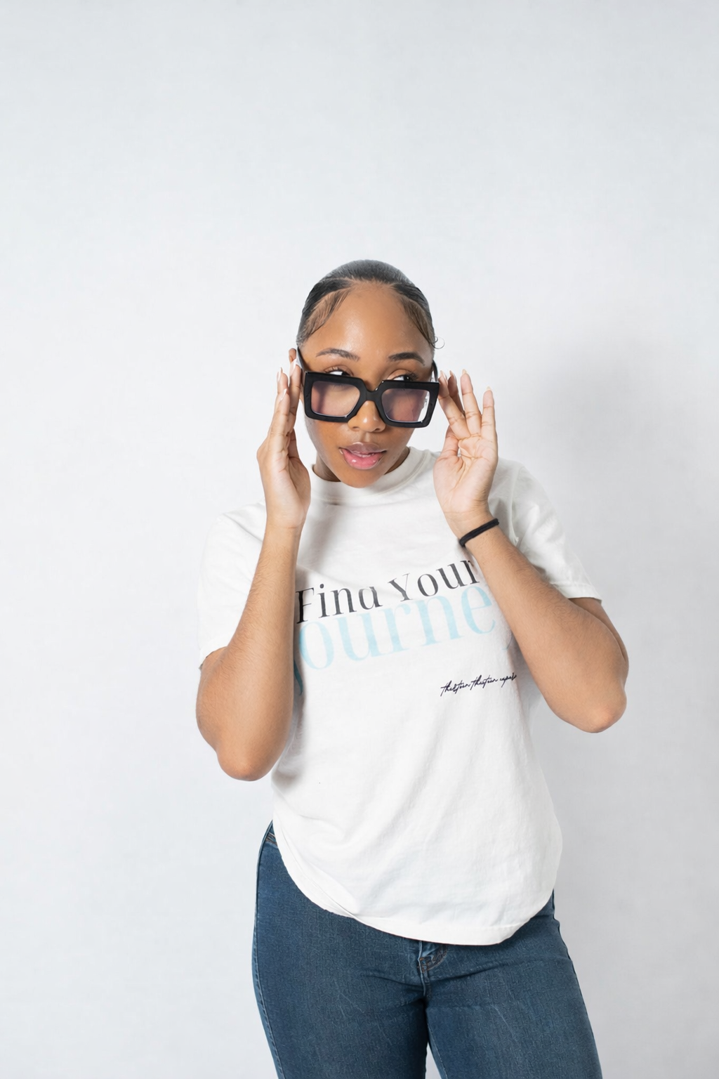 A woman wearing a white graphic t-shirt with blue and black text, dark-framed glasses, and blue jeans, posing against a light gray studio background.