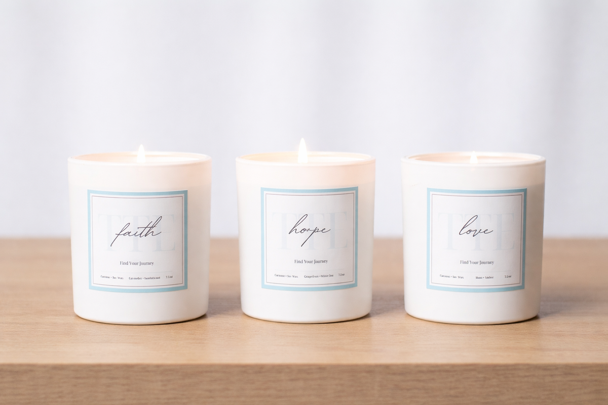 Three white glass jar candles labeled faith, hope, and love, arranged side by side on a light wood surface against a soft white background.