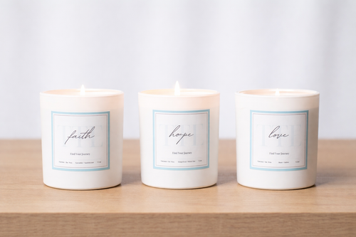 Three white glass jar candles labeled faith, hope, and love, arranged side by side on a light wood surface against a soft white background.