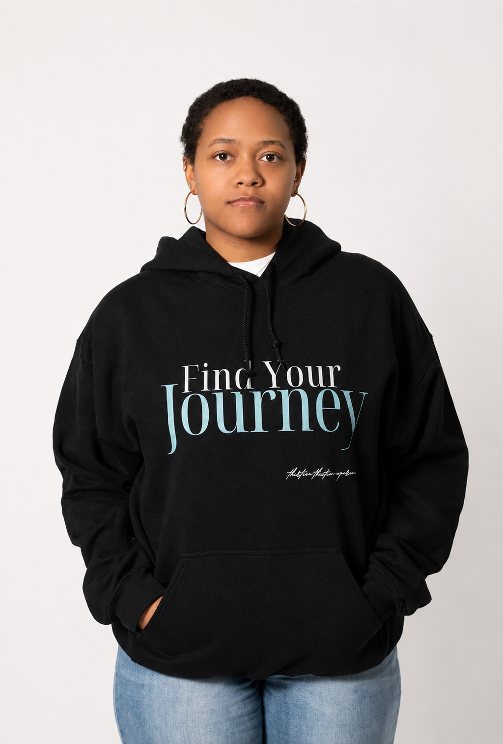 A person wearing a black pullover hoodie with the text 'Find Your Journey' printed on the chest, paired with light blue jeans, against a plain light gray background.
