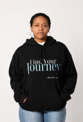 A person wearing a black pullover hoodie with the text 'Find Your Journey' printed on the chest, paired with light blue jeans, against a plain light gray background.
