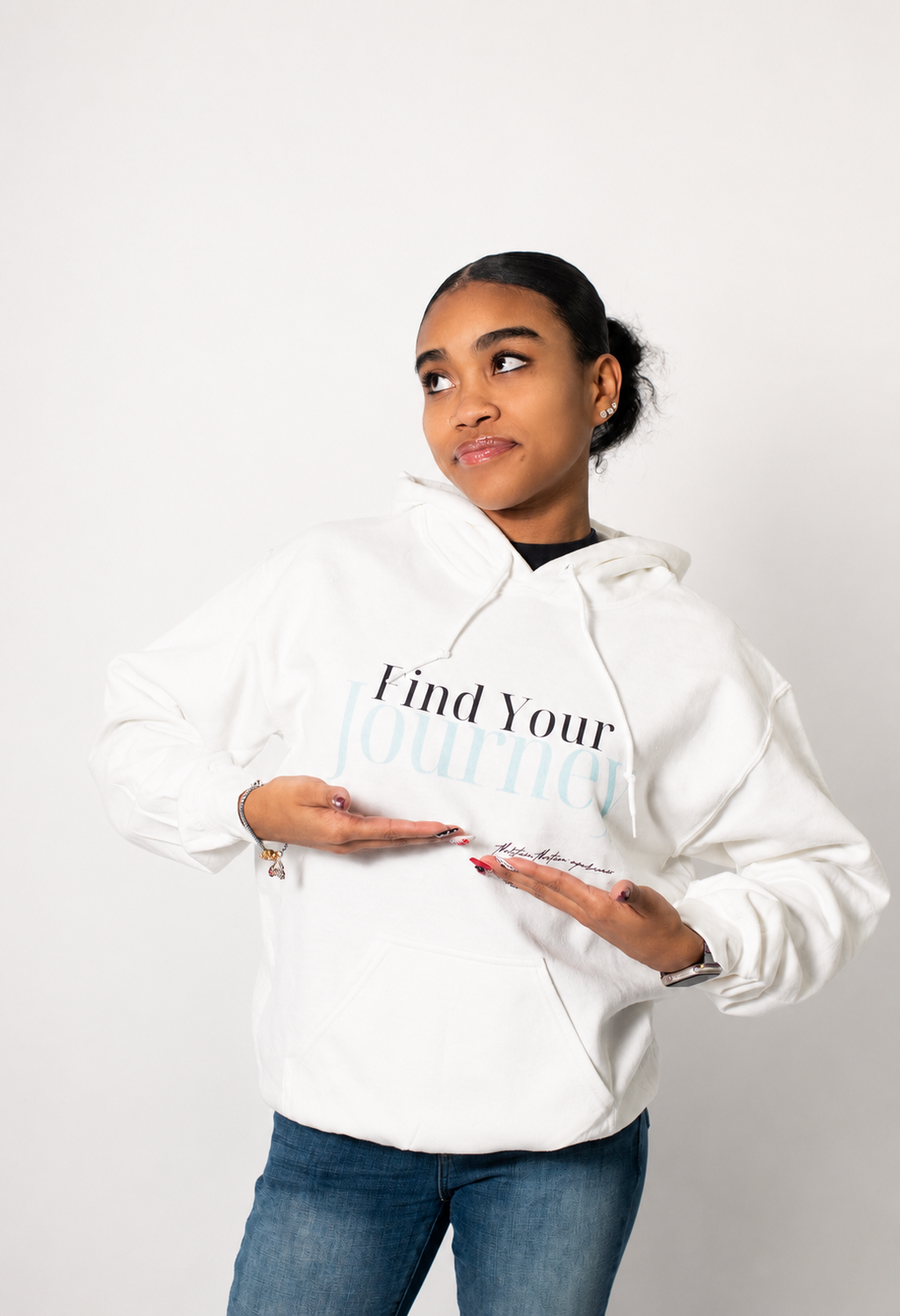 A woman wearing a white pullover hoodie with the text 'Find Your Journal' printed on the front, paired with blue jeans, standing against a plain light gray background.