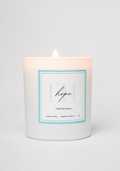 A white glass jar candle with a single lit wick, featuring a square label with a light blue border and the word 'hope' in script, set against a clean white background.